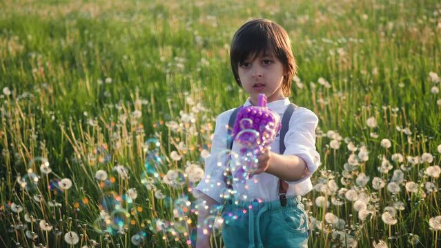 portrait of a five-year-old boy in a hat, stands on a field of dandelions and shoots soap bubbles from a toy gun at sunset in summer