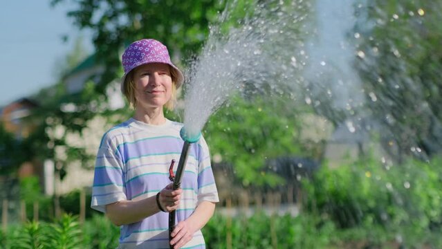 Happy female gardener watering her garden with hose in sunny summer day. Cheerful adult woman in funny hat pouring green plants in backyard.