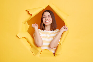Horizontal shot of smiling happy woman wearing striped shirt posing in yellow paper hole, standing with raised arms, celebrating her success, looking at camera with toothy smile.