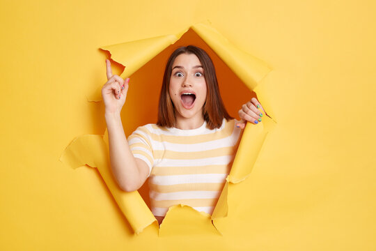 Portrait of excited beautiful woman wearing striped shirt posing in yellow paper hole, having great idea, raising finger up, keeps mouth open, looking at camera.
