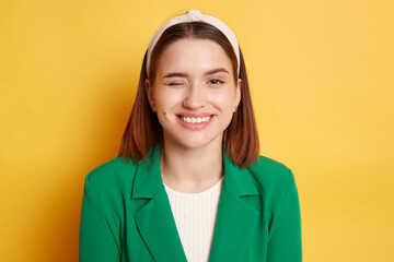 Indoor shot of playful childish woman wearing green jacket posing isolated over yellow background, looking at camera with toothy smile and winking, flirting with boyfriend.