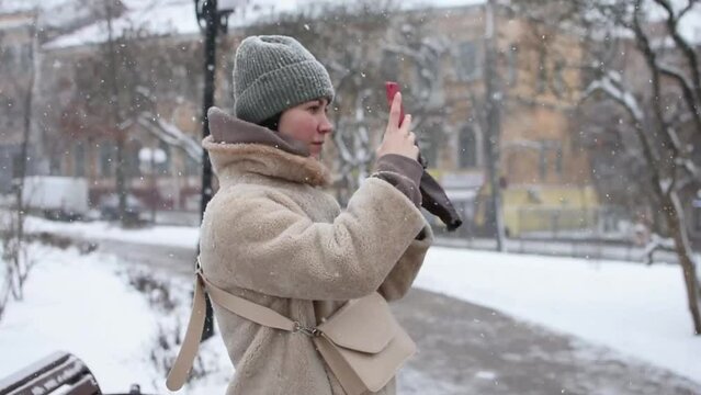 Girl in fur coat taking photo on smartphone camera on background of winter park. Snow falls, cold weather