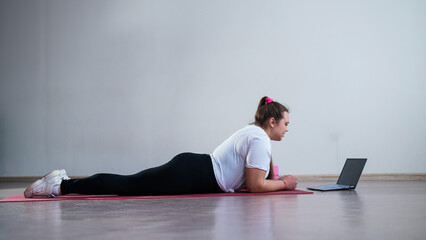 Young Plus Size Woman Stretching At Home Online. Flexible girl practices yoga and watches an online course on a laptop