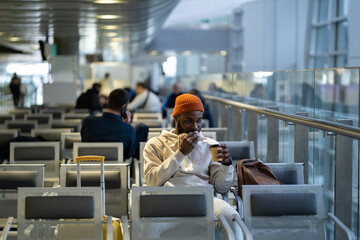 Young African American traveler man drinking coffee and eating sandwich while waiting for flying at airport terminal. Trendy black hipster guy satisfies hunger with a coffee and sandwich. 