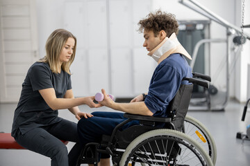 Rehabilitation specialist helps a guy to do exercises for recovery from injury, who is sitting in a wheelchair with a corset around his neck. Concept of physical therapy for people with disabilities - obrazy, fototapety, plakaty