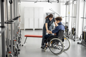 Rehabilitation specialist helps a guy stand out of a wheelchair at rehabilitation center. Concept of physical therapy and support for people with disabilities - obrazy, fototapety, plakaty