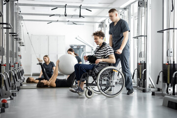 People exercising at rehabilitation center, rehabilitologist walking with guy in a wheelchair. Concept of kinesiology and recovery from injuries - obrazy, fototapety, plakaty