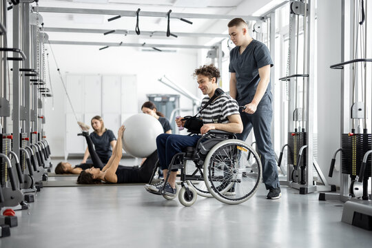 People exercising at rehabilitation center, rehabilitologist walking with guy in a wheelchair. Concept of kinesiology and recovery from injuries