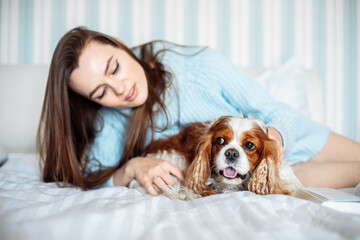 cute young woman with long hair wearing blue knitted sweater lying on bed with dog in bedroom, animal owner 