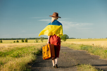 Girl in Ukraine flag with suitcase on country road in sunset