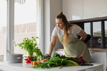 Plump, plus size blonde woman cooking healthy food in the kitchen, choosing vegetable salad, greenery. Vegetarian life 