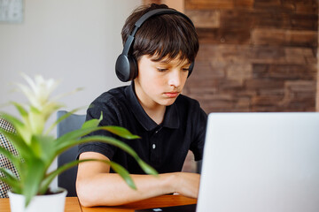 Portrait of concentrated teenager boy wearing black T-shirt, headphones sitting at desk, using...