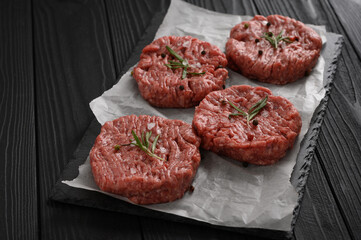 Raw beef burger patties on butcher's wooden board, rosemary and pepper. Black background. View from above.