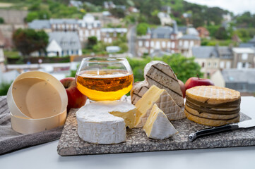 Cow cheeses of Normandy - camembert, livarot, neufchatel, pont l'eveque and glass of apple cider drink with houses of Etretat village on background, Normandy, France