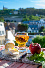 Products of Normandy, cow neufchatel lait cru cheese and glass of apple cider drink with houses of Etretat village on background, Normandy, France - obrazy, fototapety, plakaty