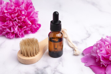 Fototapeta na wymiar Cosmetic oil in a glass dropper bottle and massage brush on marble background with peonies. Skin care cosmetic. Beauty concept for face body care