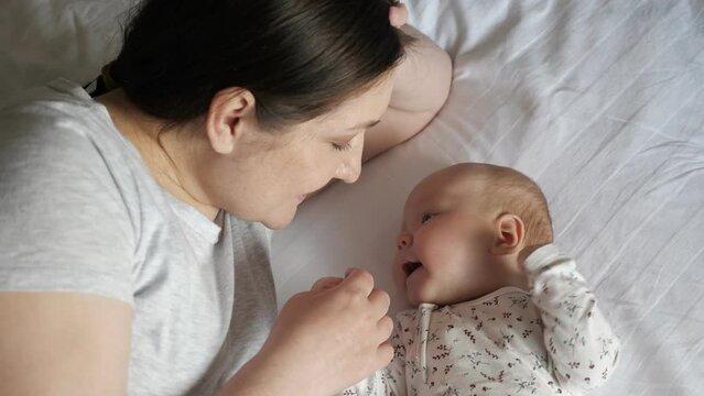 Happy mother talks to newborn daughter holding tiny hand in bedroom. Woman and adorable baby girl lie on white sheet laid on bed at hom, top view