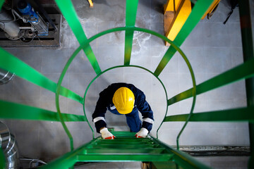 Refinery or industrial worker in safety equipment climbing up metal ladders or staircase.