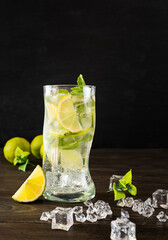 A tall transparent glass with a mojito on a dark wooden table. Slices of lime m ice cubes in the background. Black background