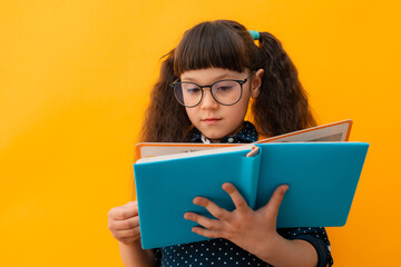 portrait of a child schoolgirl with glasses reading a book on an isolated yellow background.