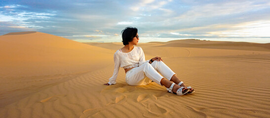short hair woman wearing sunglasses, white dress, sitting in the desert