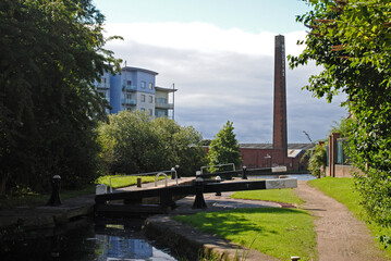 Quiet Canal with Lock Gates and Towpath with Tall Industrial Brick Chimney seen against Cloudy Sky 