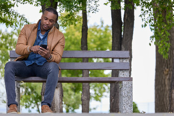 Fototapeta premium Serious young African reads a chat message from a smartphone on a bench in a city park. Stylish African-American in a brown bomber sat down on a bench to read an e-book