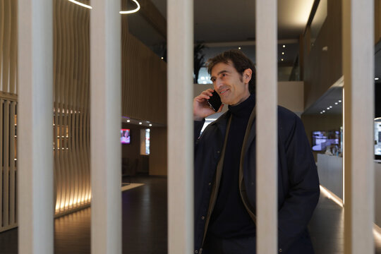 Mature businessman with smartphone by ear while entering hall of contemporary hotel and calling colleague