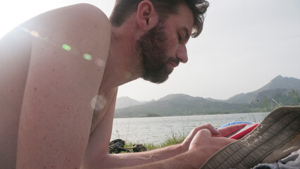 Hombre blanco barbudo sin camiseta mirando móvil en la orilla de un lago