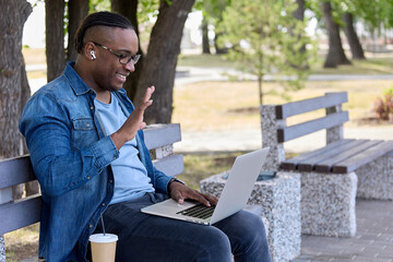 Cute guy with an African hairstyle sitting on a bench in the park asks a friend on social networks for a date. A cheerful black guy waves his hand to friends through a webcam waiting for a meeting