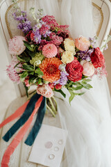 wedding bouquet with beautiful flowers on a white vintage chair against the backdrop of a veil. Next to the bouquet are wedding rings and an invitation.