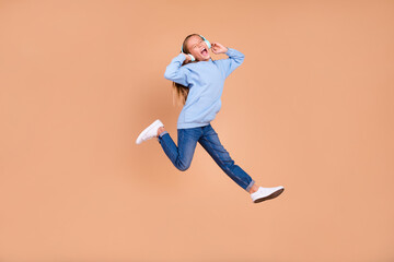 Fototapeta na wymiar Full size photo of satisfied girl jump hands touch headphones scream have fun isolated on beige color background