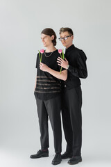 Full length of stylish gay couple holding pink tulips on grey background.