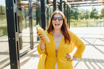 Beautiful young woman in sunglasses drinking fresh juice outdoors. Redhead girl walking in the park and holding a bottle of fresh cold smoothie in a warm sunny day. Multivitamin Juice Bottle Mockup