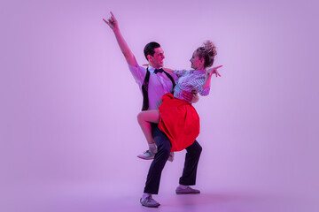 Studio shot of young man and woman in vintage retro style outfits dancing lindy hop isolated on lilac color background in neon light.