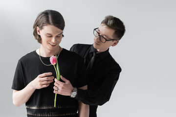 Gay in formal wear and eyeglasses holding flower near positive boyfriend isolated on grey.