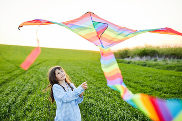 Little happy girl running with kate in hands on green wheat field. Large colored rainbow kite with long tail.
