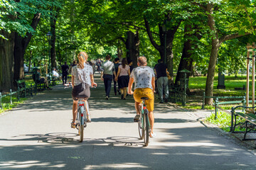 A couple of young people, a boy and a girl, ride bicycles along an asphalt path among the green trees of a city park on a sunny day, a couple walking in front of them, holding hands