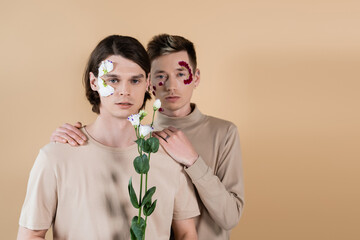Gay with petals on face hugging boyfriend with eustoma isolated on beige.