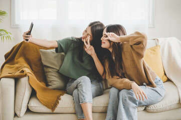 Two asian young women happy smiling and taking selfie on couch in living room at home