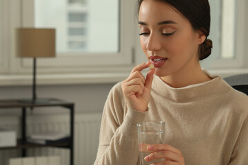 Young woman with glass of water taking dietary supplement pill indoors, space for text - obrazy, fototapety, plakaty