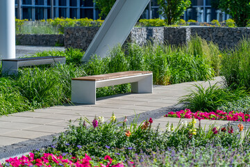 Empty concrete bench with wooden slats for sitting on tile among decorative grass and flowers in recreation area near modern office building. Garden landscape with chair in city park - obrazy, fototapety, plakaty