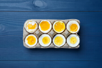 Boiled chicken eggs of different readiness stages in carton on blue wooden table, top view