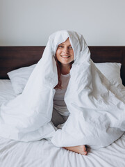 Beautiful young happy woman wrapped in soft white blanket sitting at home. Winter atmosphere. Millennial Caucasian girl wake up in cozy bed at home.