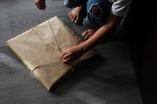 Child Unpacks A Gift. Kid Hands Untie The Rope. Craft Paper Present Unpacking