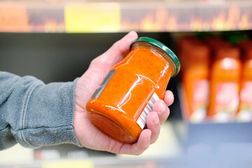 male hand holds jar of sauce on blurred background, row of shelves with groceries in supermarket,...
