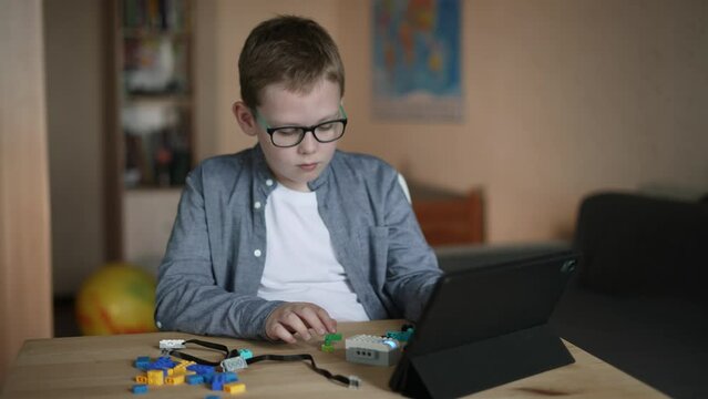 Caucasian elementary school boy in glasses sits at table with tablet, collects details of plastic constructor with electronic elements with smile. Child connects parts of his own product with effort.