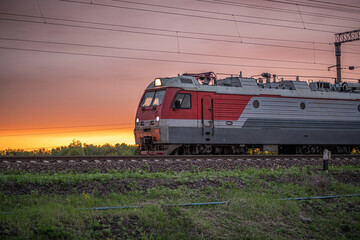 The railroad train locomotive of the gray-red train rushes forward at dawn on a summer morning among low green grass on sunrise or sunset
