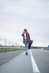 Fototapeta premium Cool woman with curly brown hair, in an unbuttoned plaid shirt, torn jeans, a white turtleneck, high heels, sitting on the road fence with electric guitar. Hitchhiker or traveler musician