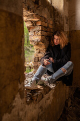 A beautiful woman with curly brown hair in rock Gothic style: torn jeans, black mantle, sitting on the broken window of an abandoned industrial building with brick walls. Post apocalypse danger world